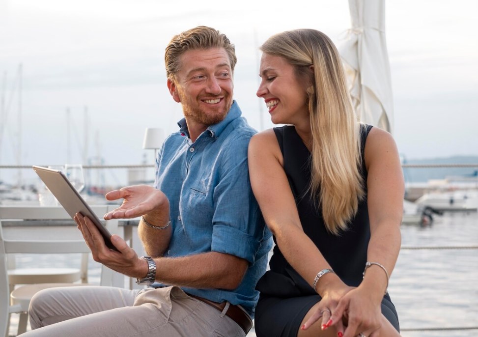 a smiling couple using a tablet on a yacht, with other yachts in the background